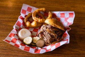 Dyer's BBQ pulled pork sandwich with onions and pickles, onion rings on the side plated in a basket with red and white checkered paper